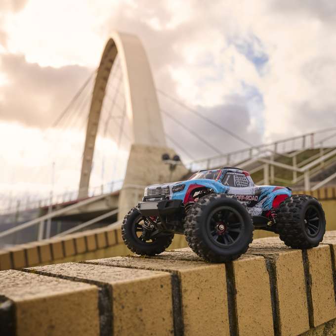 1:16 mēroga RC automašīna ar bezsuku motoru un pilnpiedziņu (4WD), izvietota uz ķieģeļu sienas ar miglainu fonu, parādot izcilu dizainu un izturību
