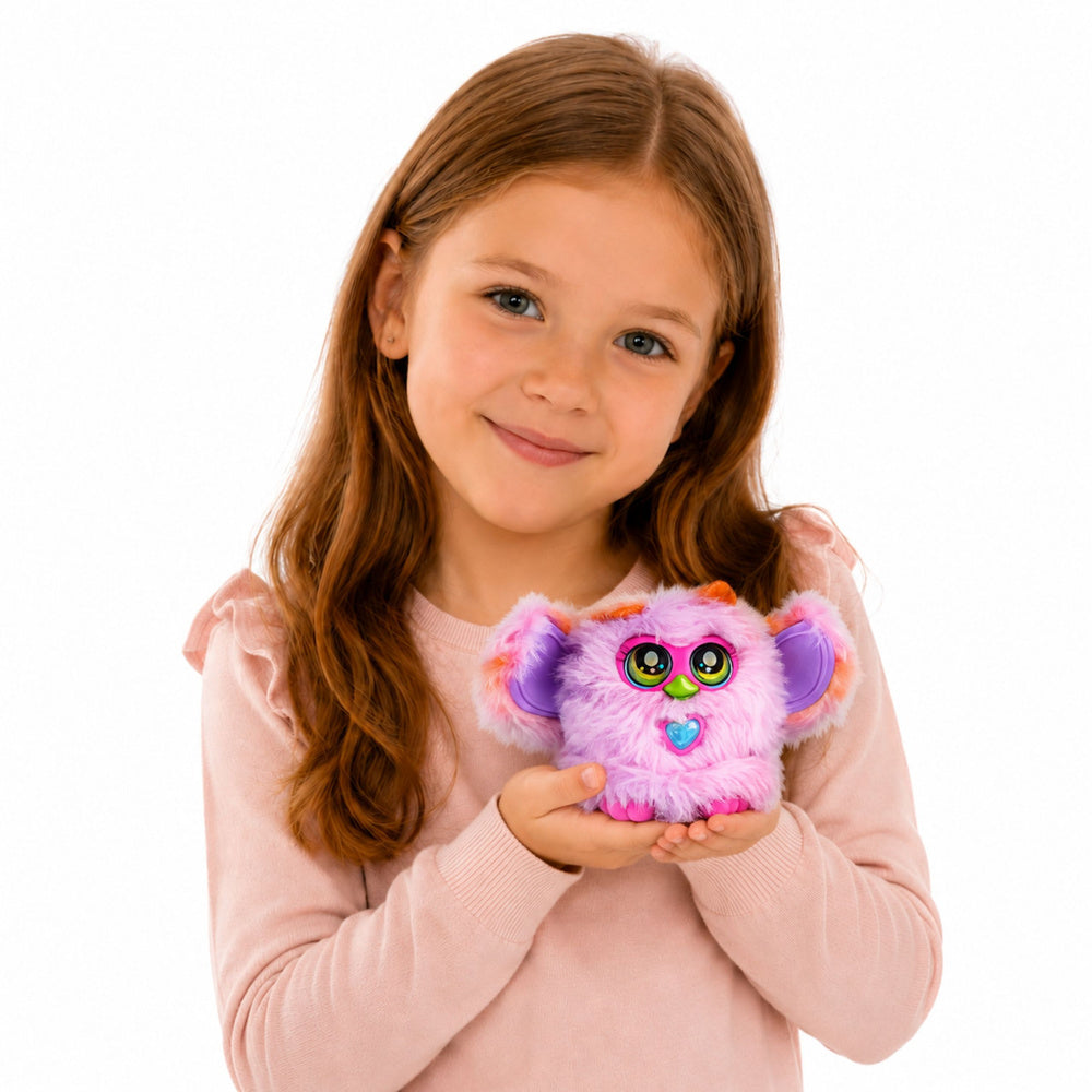 Smaidīga meitene rozā džemperī tur rokās interaktīvo pūkaino L-Toys Genie Pets briesmonīti violetā un rozā krāsā ar lielām acīm, kustīgām ausīm un sirsniņas formas pogu