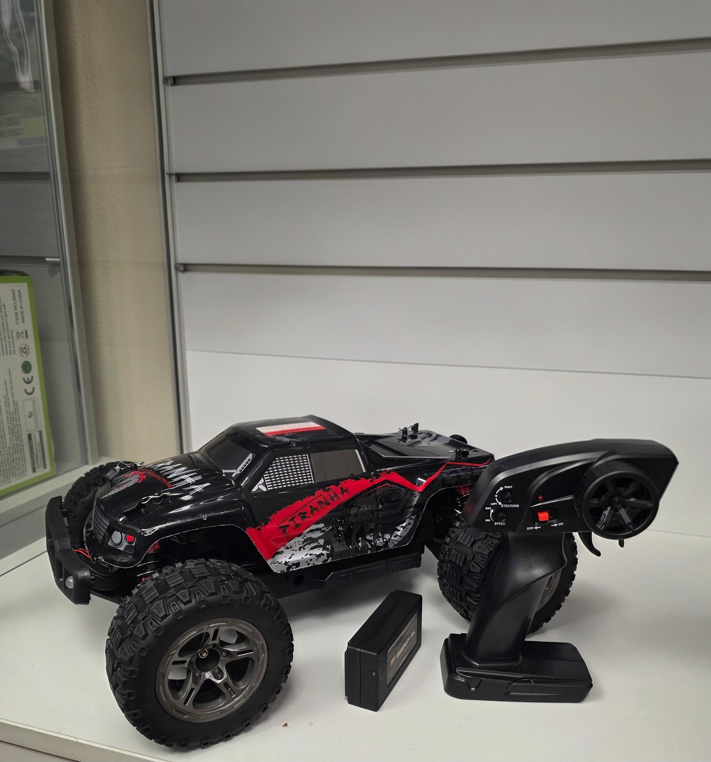 Melns RC monstrtrača modelis ar lieliem riteņiem, tālvadības pulti un akumulatoru uz baltas virsmas.