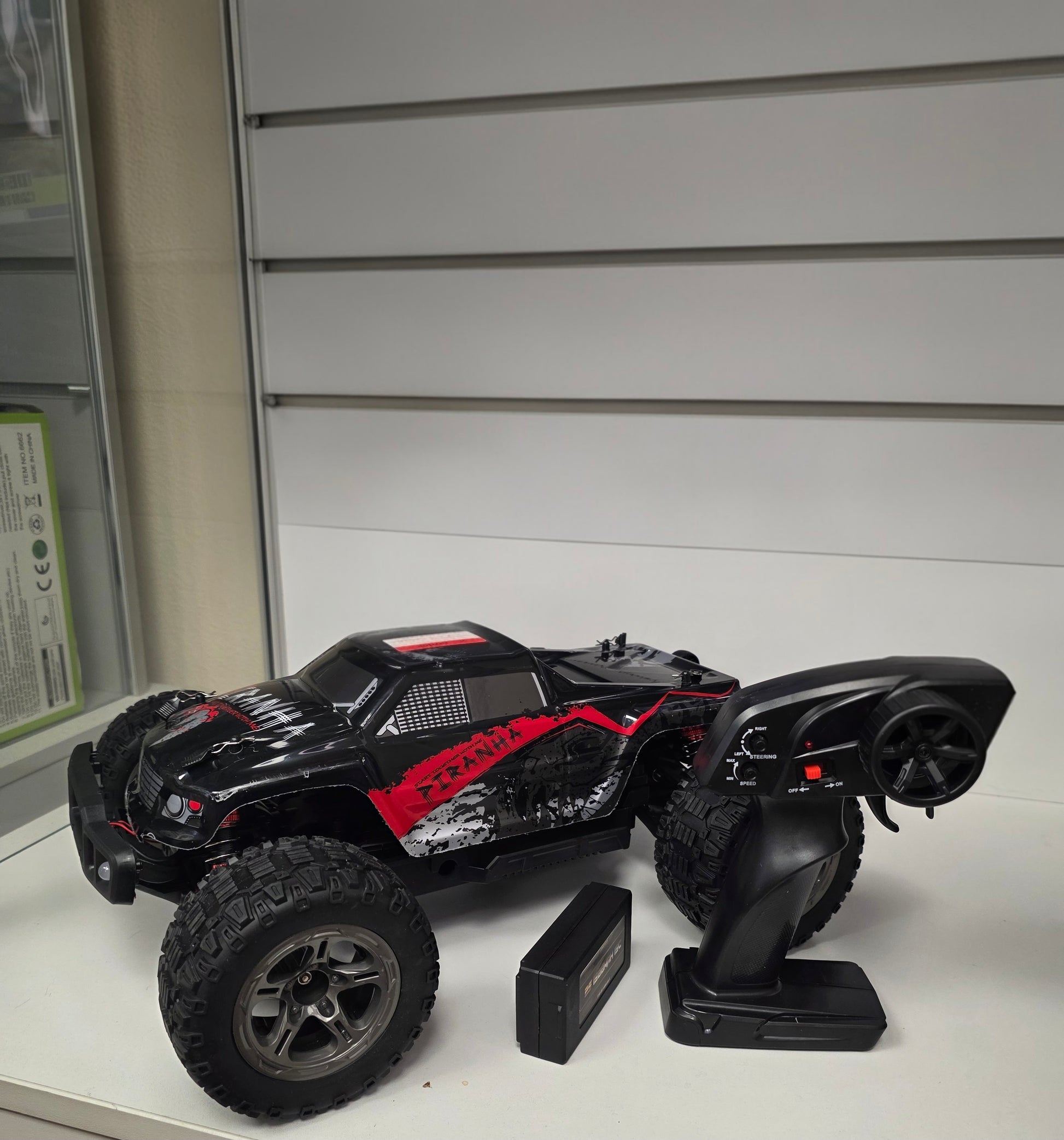 Melns RC monstrtrača modelis ar lieliem riteņiem, tālvadības pulti un akumulatoru uz baltas virsmas.
