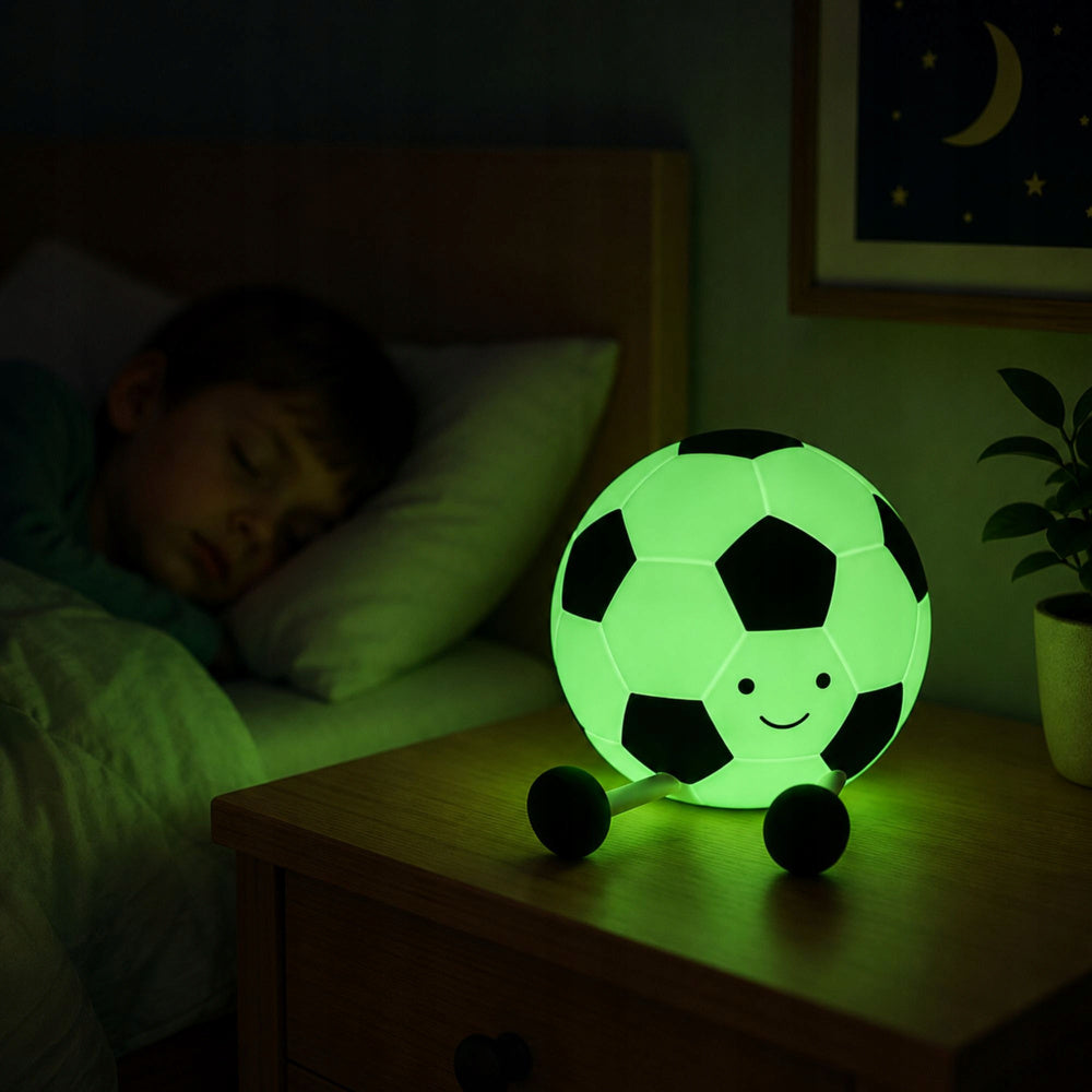 Mīksta silikona futbola bumbas LED RGB naktslampiņa zaļā gaismā uz naktsgaldiņa blakus guļošam bērnam